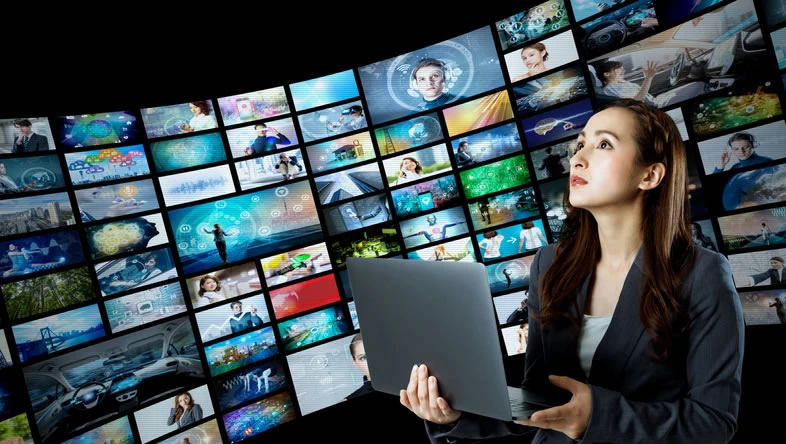 woman holding laptop surrounded by many video screens